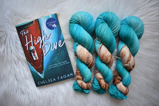 Book titled 'The High Dive' by Chelsea Fagan next to skeins of blue yarn with brown accents on a white fluffy surface.